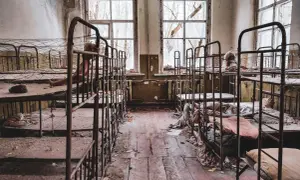 Opuszczony pokój z rzędami zardzewiałych łóżek piętrowych, zniszczonymi oknami i śmieciami na podłodze.