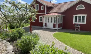 Ett rött hus med vita detaljer och en vacker farstukvist som leder in till hallen. En blommande äppelträd och grönskande trädgård.