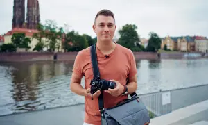 Mężczyzna z aparatem w rękach, w plenerze nad rzeką, z widokiem na zabytkowe budynki i katedrę.