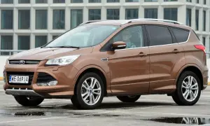 Brązowy Ford Kuga 2 z kierowcą w okularach i kratę, zaparkowany na mokrym parkingu przed nowoczesnym budynkiem.