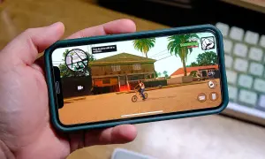 Ręka trzyma smartfon z grą RPG na telefon. Na ekranie widać postać na rowerze przed domem, palmy i ikony sterowania.