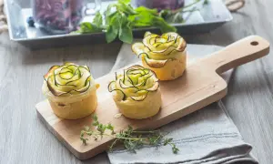 Trois petites bouchées en forme de rose, ornées de fines lamelles de fleur de courgette sur une planche en bois.