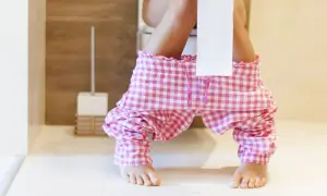 Kobieta w różowych spodniach w kratkę siedzi na toalecie, obok stoi szczotka. Czas na poranną toaletę.
