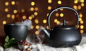 Foto still life: una teiera di ghisa nera emana vapore, accanto a una ciotola fumante e a ingredienti per il tè su uno sfondo di luci soffuse.