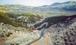 Vue aérienne d'une route sinueuse traversant un paysage montagneux verdoyant, menant à un village sur le plateau de Lassithi. Des ruines de moulins à vent parsèment la colline.