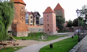 Chojnice atrakcje dla dzieci: plac zabaw przy zabytkowych wieżach. Dzieci bawią się w piaskownicy i na huśtawkach, a w tle widać ceglane budowle.