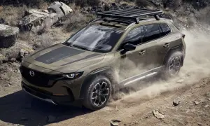 Brązowa Mazda CX-50 z bagażnikiem dachowym przemierza pustynny teren, wzbijając tumany kurzu.