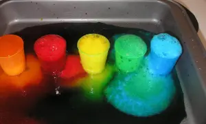 Kolorowe kubeczki z pianą w tacy, tworzące tęczę z płynów.
