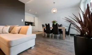 Nowoczesny salon z jadalnią i aneksem kuchennym. Jasna kanapa, drewniany stół z krzesłami, wysoka minimalna wysokość pomieszczeń.