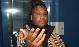 A young man with dreadlocks, wearing a plaid jacket, gestures forward. He's the artist behind many popular Tay Keith songs.
