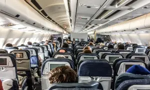 Blick in den Gang eines Flugzeugs. Viele Passagiere sitzen auf ihren Plätzen im Flugzeug innen, einige schauen auf die Bildschirme vor sich.