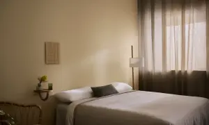 Ett lugnt sovrum med en inbjudande säng, en golvlampa och en liten hylla med en vas och en blomma.