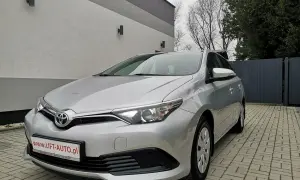Srebrna Toyota Auris 1.6 benzyna zaparkowana na podjeździe z kostki brukowej, przed nowoczesnym budynkiem.