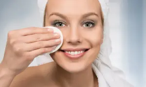 Donna sorridente con asciugamano in testa, pulisce il viso con un dischetto. Scopri i rimedi della nonna per pori dilatati.