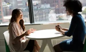 Dos mujeres conversan animadamente en una oficina, con una vista urbana de fondo. Una de ellas gesticula mientras habla, demostrando un nivel de **speaking B2** en su comunicación.