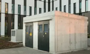 Betonowa stacja transformatorowa z dwoma czarnymi drzwiami i żółtymi znakami ostrzegawczymi. W tle nowoczesny budynek z białymi i ciemnymi panelami.