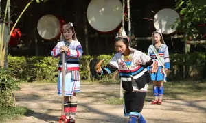 Trzy dziewczyny w barwnych strojach ludowych, jedna trzyma kij, druga instrument, w tle wielkie bębny.