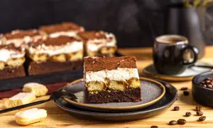 Ein Stück Tiramisu Brownie mit Löffelbiskuits, cremiger Füllung und Kakaopulver. Daneben eine Tasse Kaffee und Kaffeebohnen.