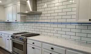 White subway tile backsplash with green grout, a stainless steel range hood, and white cabinets. This image helps visualize the cost to install tile backsplash.