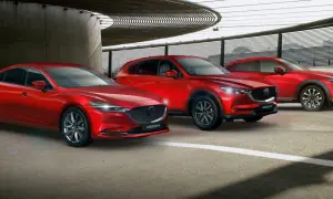 Trzy czerwone samochody marki Mazda: sedan, SUV i crossover, zaparkowane pod nowoczesną konstrukcją.