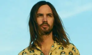 Kevin Parker w koszuli z motywem oczu, z rozwianymi włosami na tle błękitnego nieba.