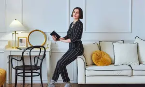 Elegancka kobieta w paski, w stylu glamour ubrania, siedzi na sofie, trzymając książkę. Obok biurko z lampą i lustrem.