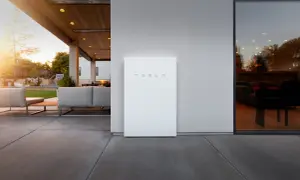 Un elegante Tesla Powerwall blanco se alza junto a una casa moderna con un patio y muebles de exterior.