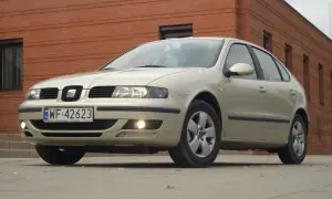 Jasny Seat Leon 1 z włączonymi światłami mijania i przeciwmgielnymi, zaparkowany przed ceglanym budynkiem.
