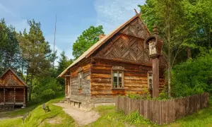 Drewniane chaty i kapliczka na słupie w skansenie w Polsce. W tle las i błękitne niebo.