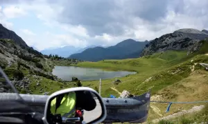 Widok z motocykla na malownicze **przełęcze alpejskie**, jezioro i góry pod zachmurzonym niebem.