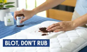 A person blots a stain on a white mattress topper with a cloth. The mattress topper is on a blue mattress. The text "BLOT, DON'T RUB" is visible.