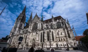 Der imposante Dom Regensburg ragt mit seinen Türmen in den blauen Himmel. Menschen und Autos bewegen sich auf dem Platz davor.