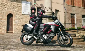 Para jadąca motocyklem po brukowanej uliczce, oboje w kaskach i kombinezonach.