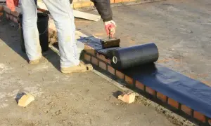 Pracownik kładzie hydroizolację na murze, przygotowując zbrojenie pod dalsze prace budowlane.