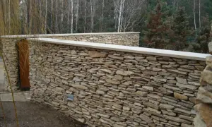Solidne ogrodzenie z piaskowca, z drewnianymi drzwiami i kamiennymi lampkami, otacza ogród.