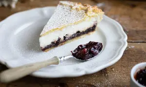 Una fetta di crostata ricotta e visciole, cosparsa di zucchero a velo, su un piatto bianco. Un cucchiaino è pieno di visciole sciroppate.