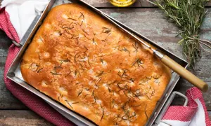 En gyllene focaccia rosmarin i en plåt, toppad med örter och flingsalt. En kniv ligger på brödet, redo att skäras.