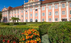 Das Meersburg Neues Schloss erstrahlt in Rosa vor blauem Himmel. Ein gepflegter Garten mit bunten Blumen und Palmen lädt zum Verweilen ein.