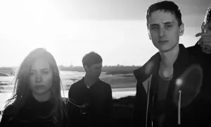 Cztery osoby na tle zachodzącego słońca. W tle widać wodę i trawę. To okładka albumu Arctic Monkeys "Suck It and See".