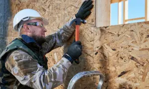 Budowlaniec w kasku i okularach zabezpieczających przybija młotkiem płytę OSB. Impregnat do płyty OSB zapewni jej trwałość.