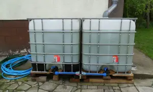 Dwa zbiorniki na deszczówkę połączone rurą, gotowe do podłączenia do rynny. Wąż ogrodowy leży obok.