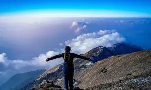 Eine Frau steht auf einem Berggipfel, die Arme ausgebreitet, und blickt auf eine Wolkendecke. Die Frage "wie hoch ist der Kilimandscharo?" hallt in der Weite wider.