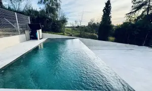 Una moderna piscina infinita con agua turquesa, rodeada de un área de concreto blanco y vegetación exuberante. El cielo azul con nubes blancas completa la escena.