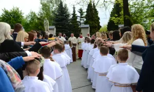 Dzieci w białych strojach komunijnych stoją w rzędzie, a dorośli kładą im ręce na głowach.