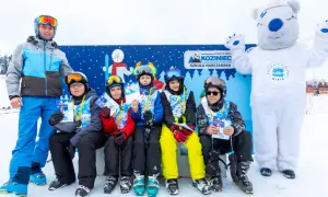 Uśmiechnięci młodzi narciarze z dyplomami i medalami po zajęciach w szkole narciarskiej Koziniec. Towarzyszy im instruktor i biały miś.