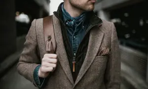 Mann im Tweed-Sakko mit Jeanshemd und Halstuch, perfekt für den party chic dress code male. Braune Ledertasche mit Coach-Anhänger.