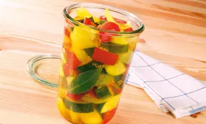 Ein Glas mit bunter Mischung aus Zucchini, Paprika und Tomaten, bereit zum Zucchini einkochen. Senfkörner und Lorbeerblätter sind ebenfalls sichtbar.