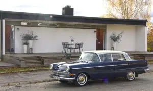 En mörkblå Opel Kapitän från 1960 står parkerad framför ett modernt hus med träaltan.