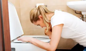 Kobieta w białej koszulce pochyla się nad toaletą, doświadczając silnych wymiotów.