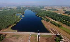 Widok z lotu ptaka na zalew Jagodno otoczony lasem i polami. Woda lśni w słońcu, a tama z budowlami hydrotechnicznymi stanowi imponującą konstrukcję.